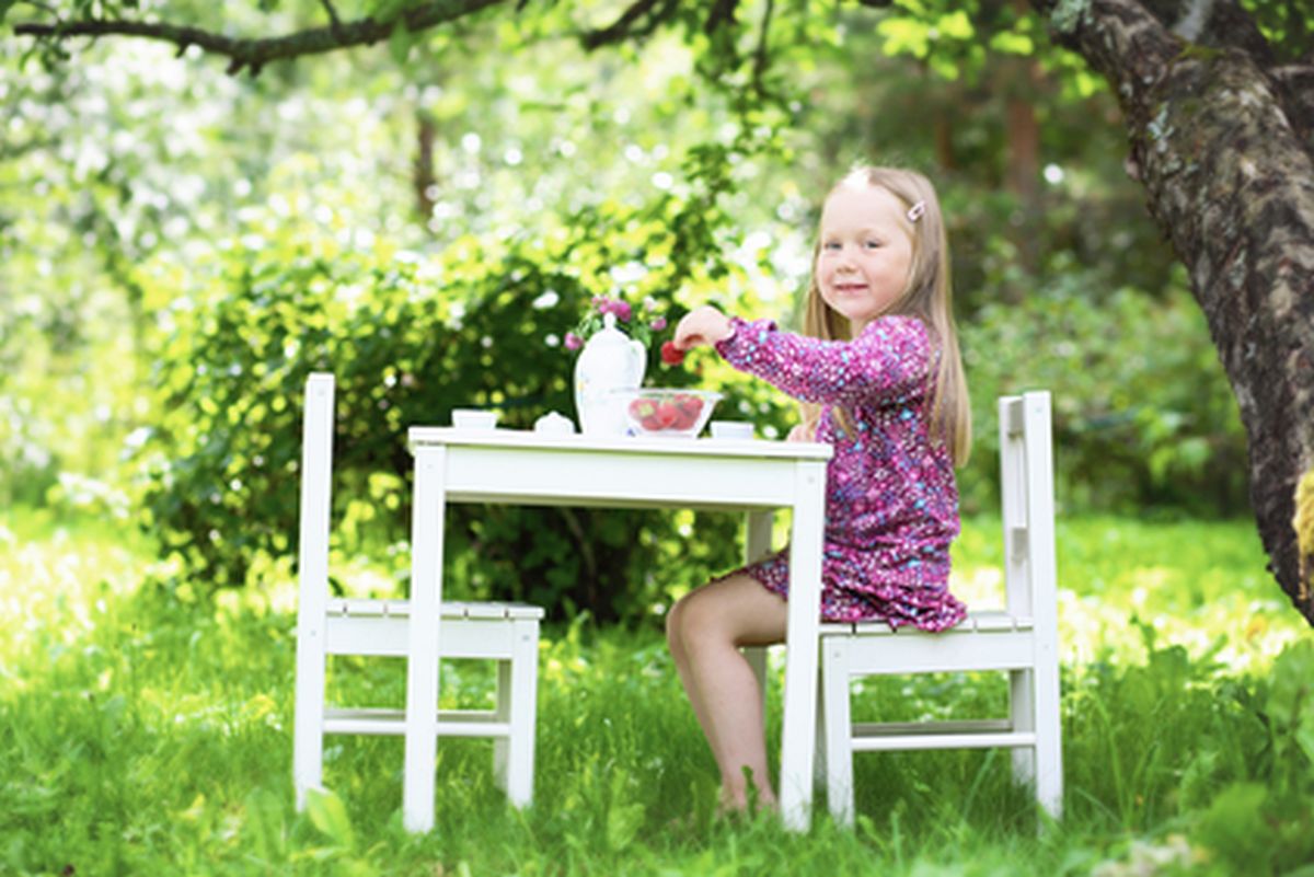 A smiling little girl sitting at a white table having a tea party in a garden setting on a sunny summer day while holding a delicious strawberry.
