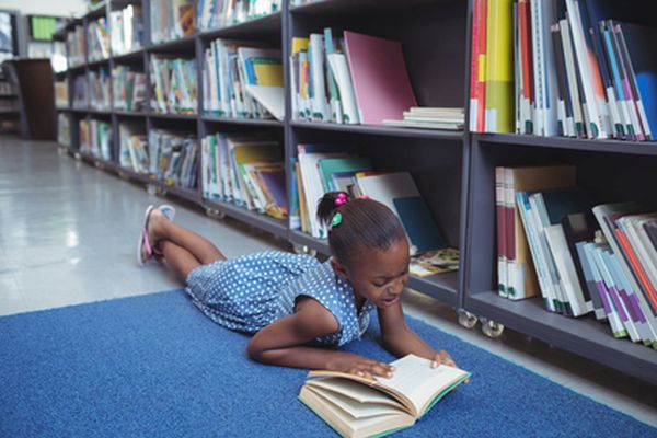 Girl reading book while lying by shelf in library